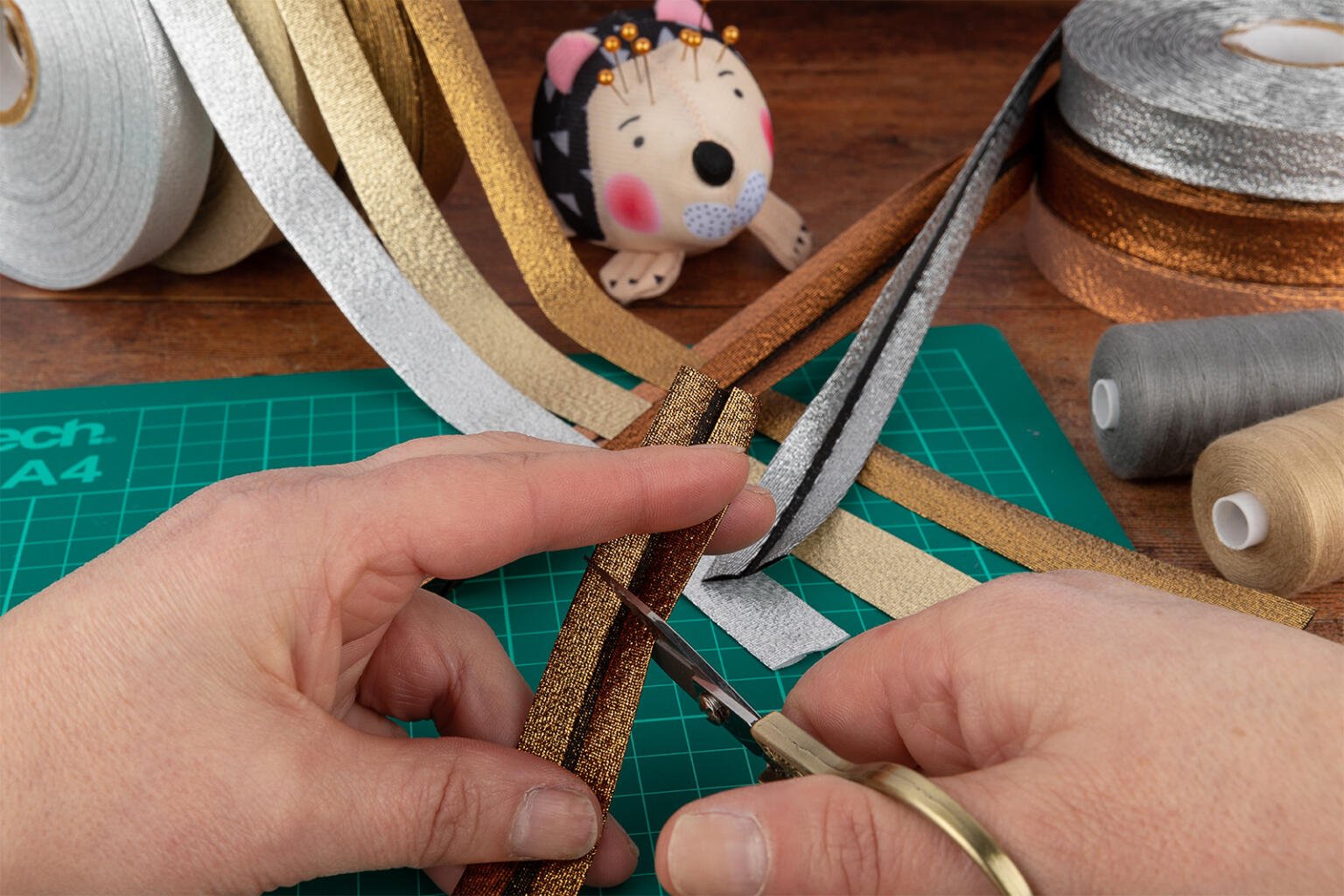 Close-up of hands cutting gold glitter ribbon with scissors on a green cutting mat. Surrounding the hands are various rolls of metallic ribbon, gray thread bobbins, and a fabric pincushion shaped like an animal with pins on its back.