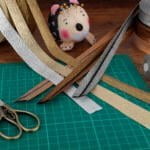 A crafting table with various supplies: gold, silver, bronze, and black glittery ribbons, a pair of vintage-style scissors, a green self-healing cutting mat, spools of gray and beige thread, and a hedgehog-shaped pincushion with pins.