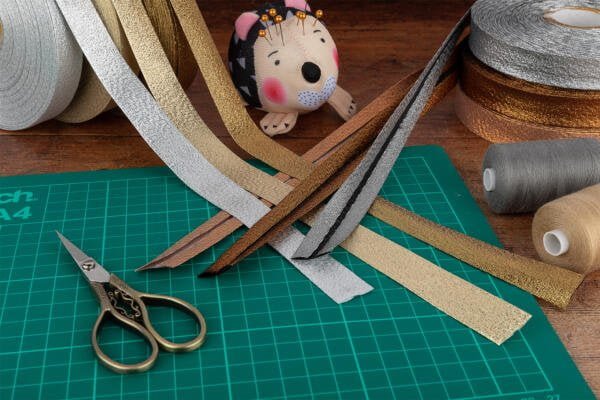A crafting table with various supplies: gold, silver, bronze, and black glittery ribbons, a pair of vintage-style scissors, a green self-healing cutting mat, spools of gray and beige thread, and a hedgehog-shaped pincushion with pins.