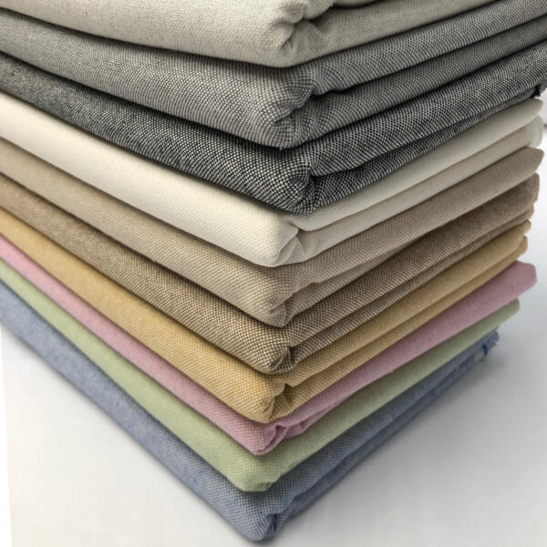 A stack of neatly folded, solid-colored fabric swatches arranged in a gradient from dark to light. The fabrics include various shades of gray, beige, cream, tan, pink, lavender, green, and blue, showcasing a range of pastel and neutral tones.