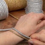 Close-up of a person wrapping silver and gold metallic cords around their wrist as if trying on bracelets. Spools of the same cords are visible in the background on a wooden table. The person's hands are prominently featured, demonstrating the craft activity.