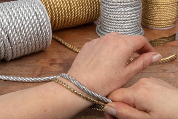 Close-up of a person wrapping silver and gold metallic cords around their wrist as if trying on bracelets. Spools of the same cords are visible in the background on a wooden table. The person's hands are prominently featured, demonstrating the craft activity.