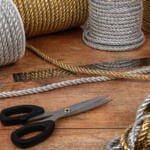 Coils of thick, twisted silver and gold rope are arranged on a wooden surface along with a pair of black-handled scissors. One rope section is cut in the middle, and several of the ropes are partially unwound, showing their textured fibers.