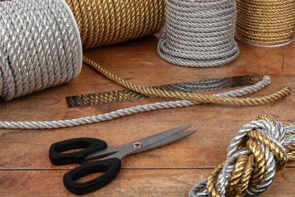 Coils of thick, twisted silver and gold rope are arranged on a wooden surface along with a pair of black-handled scissors. One rope section is cut in the middle, and several of the ropes are partially unwound, showing their textured fibers.