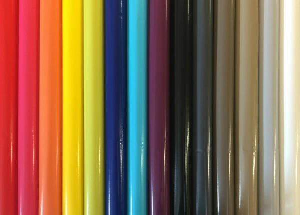 A close-up image of colorful rolls of vinyl arranged vertically. The colors, from left to right, include bright red, orange, light orange, yellow, bright yellow, blue, cyan, turquoise, purple, black, dark grey, grey, light grey, and white.