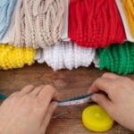 A person's hands holding a measuring tape against a teal-colored yarn on a wooden surface. Around them are bundles of colorful yarn in shades of beige, red, yellow, white, green, teal, and lemon yellow. A yellow tape measure and sewing pins are also visible.