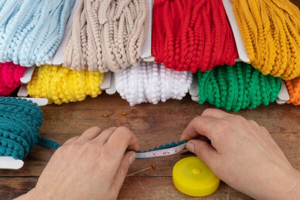 A person's hands holding a measuring tape against a teal-colored yarn on a wooden surface. Around them are bundles of colorful yarn in shades of beige, red, yellow, white, green, teal, and lemon yellow. A yellow tape measure and sewing pins are also visible.