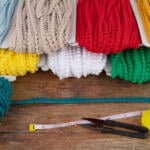 A variety of colorful yarns in different textures and shades, including red, yellow, green, white, and blue, are neatly arranged on a wooden surface. In front, there is a tape measure, scissors, and a seam ripper with golden pins scattered around.