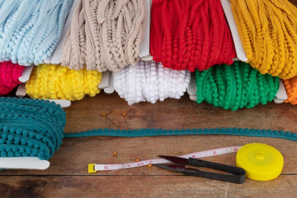 A variety of colorful yarns in different textures and shades, including red, yellow, green, white, and blue, are neatly arranged on a wooden surface. In front, there is a tape measure, scissors, and a seam ripper with golden pins scattered around.
