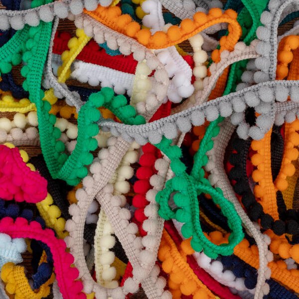 A close-up image of various colorful pom pom trims intertwined in a chaotic arrangement. The trims feature small, round pom poms in shades of red, green, yellow, blue, orange, white, and gray, creating a vibrant and textured pattern.
