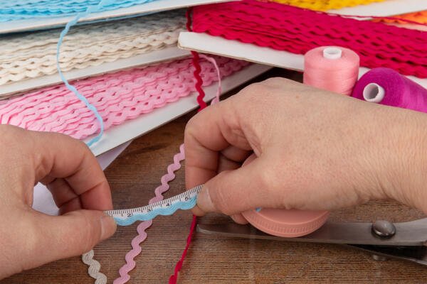 Close-up of hands measuring light blue rickrack with a small tape measure, surrounded by spools of pink and red thread, scissors, and various colors of rickrack coils in the background. The scene is set on a wooden table, suggesting a sewing or crafting project.