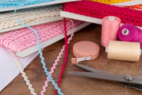 A wooden table with various sewing supplies, including spools of thread in pink, purple, and beige, folded trims in different colors and patterns, a pink measuring tape, and a pair of scissors. The scene suggests a crafting or sewing project in progress.
