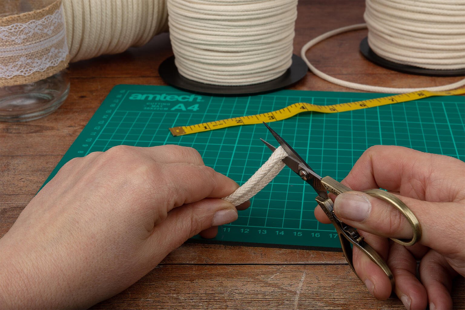 Two hands cut a piece of beige rope with small scissors on a green grid cutting mat. A yellow measuring tape, spools of lace and cord, and a glass jar are visible in the background on a wooden surface.