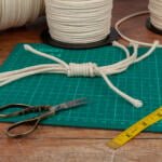 A green cutting mat with a knotted bundle of rope on it. Scissors, a yellow measuring tape, and rolls of rope are placed around the mat on a wooden table. A glass jar with lace and additional tools is also partially visible.