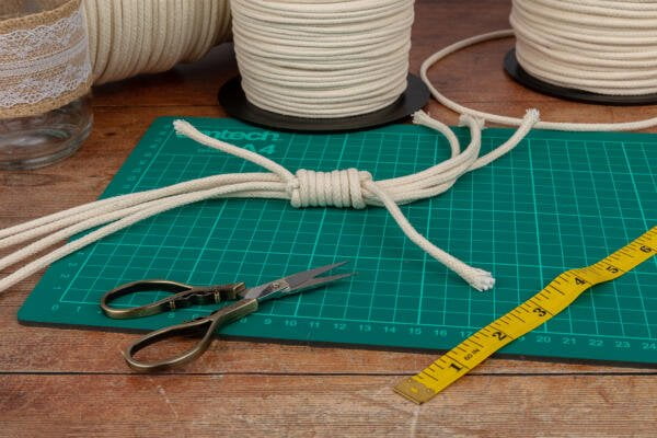 A green cutting mat with a knotted bundle of rope on it. Scissors, a yellow measuring tape, and rolls of rope are placed around the mat on a wooden table. A glass jar with lace and additional tools is also partially visible.