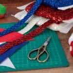 A crafting workspace with colorful materials: rolls of red, blue, and white textured trims, fabric sheets, a pair of scissors, spools of red and white thread, and a strawberry-shaped pincushion with pins on a green cutting mat on a wooden table.