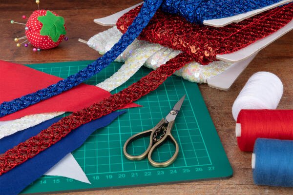 A crafting workspace with colorful materials: rolls of red, blue, and white textured trims, fabric sheets, a pair of scissors, spools of red and white thread, and a strawberry-shaped pincushion with pins on a green cutting mat on a wooden table.