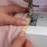 Hands guiding a piece of pink fabric with lace trim under the needle of a white sewing machine. The sewing machine is stitching the lace trim to the fabric.