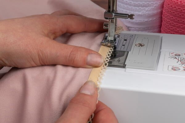 Hands guiding a piece of pink fabric with lace trim under the needle of a white sewing machine. The sewing machine is stitching the lace trim to the fabric.