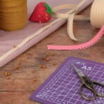 A crafting workspace with lace ribbons, pin cushions, yellow-headed pins, scissors, and a cutting mat on a wooden surface. The lace ribbons are in various colors, including red, beige, and pink. A strawberry-shaped pin cushion is also visible.
