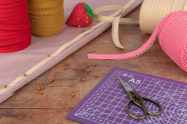 A crafting workspace with lace ribbons, pin cushions, yellow-headed pins, scissors, and a cutting mat on a wooden surface. The lace ribbons are in various colors, including red, beige, and pink. A strawberry-shaped pin cushion is also visible.