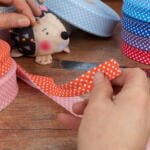 Hands are holding scissors and cutting a piece of red and white polka dot ribbon on a wooden surface. A pin cushion shaped like an animal and various rolls of polka dot ribbon in different colors are visible in the background, along with a metal ruler.