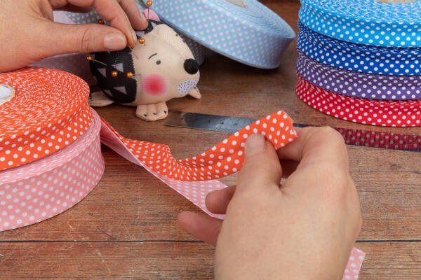 Hands are holding scissors and cutting a piece of red and white polka dot ribbon on a wooden surface. A pin cushion shaped like an animal and various rolls of polka dot ribbon in different colors are visible in the background, along with a metal ruler.