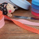 A sewing scene with rolls of polka-dotted ribbon in various colors, including blue, pink, red, and purple, scattered on a wooden surface. A metal ruler lies flat, and a pin cushion in the shape of a hedgehog is placed among the ribbons.