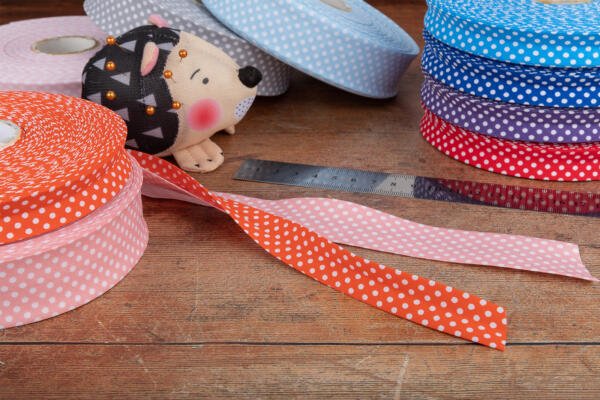 A sewing scene with rolls of polka-dotted ribbon in various colors, including blue, pink, red, and purple, scattered on a wooden surface. A metal ruler lies flat, and a pin cushion in the shape of a hedgehog is placed among the ribbons.
