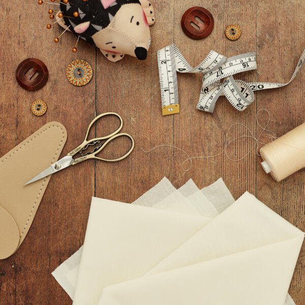 A flat lay of sewing supplies on a wooden surface. Items include beige fabric, a tape measure, spools of thread, a pair of scissors in a leather case, buttons, and a pin cushion shaped like a hedgehog with various pins stuck in it.