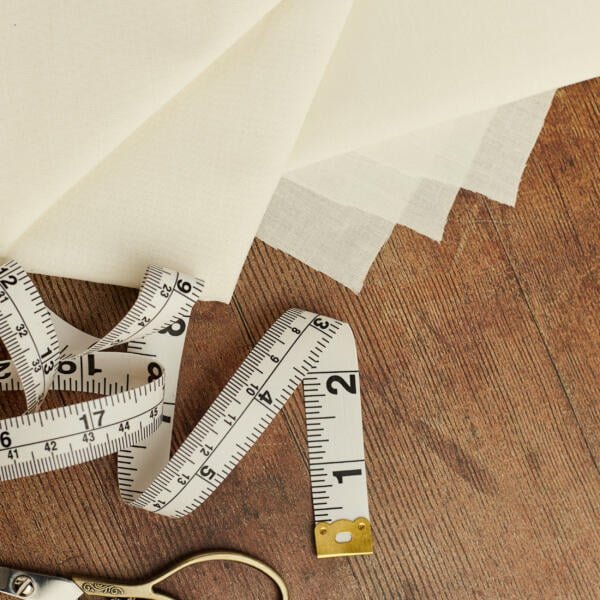 A white cloth and a few sheets of white fabric are laid out on a wooden surface. A white measuring tape is draped over the fabric, and a pair of silver scissors is partially visible at the bottom left corner.