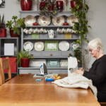 An elderly woman with short, white hair is sewing fabric at a large wooden table in a cozy room. The background features a cabinet with decorative plates, plants, and mugs. The table holds sewing supplies, including a sewing machine, an iron, and fabric.