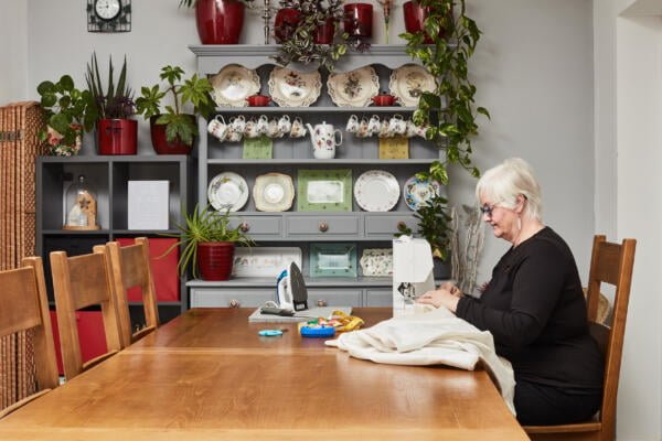 An elderly woman with short, white hair is sewing fabric at a large wooden table in a cozy room. The background features a cabinet with decorative plates, plants, and mugs. The table holds sewing supplies, including a sewing machine, an iron, and fabric.