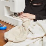 A person in a black top uses a white sewing machine to stitch fabric on a wooden table. A blue pincushion with colorful pins is placed nearby. The scene suggests they are engaged in sewing or tailoring work.