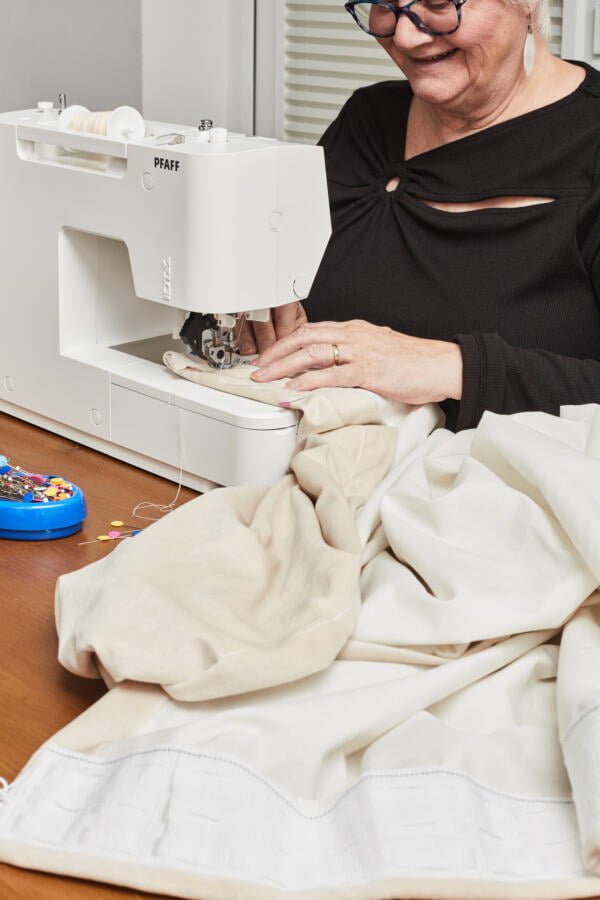 A person in a black top uses a white sewing machine to stitch fabric on a wooden table. A blue pincushion with colorful pins is placed nearby. The scene suggests they are engaged in sewing or tailoring work.