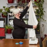 An elderly woman with short white hair and glasses is standing next to a sewing machine, holding a piece of white fabric. She is in a room with potted plants, a shelf with plates, and a sign that reads "Reserved for Dad." A pin cushion and blue scissors are on the table.