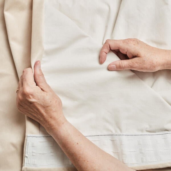 Close-up of two hands examining fabric. One hand holds a corner of a white piece of fabric, while the other hand holds onto a beige fabric beneath. The background shows more of the same white and beige fabric. The image highlights the texture and layer of the fabrics.