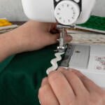 Close-up of hands guiding green fabric with white ric-rac trim through a sewing machine. The machine is actively stitching, with the needle down on the trim. Various colors of ric-rac trim are visible in the background.