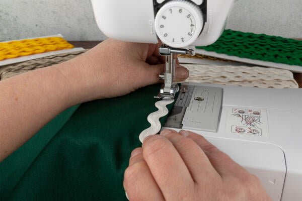 Close-up of hands guiding green fabric with white ric-rac trim through a sewing machine. The machine is actively stitching, with the needle down on the trim. Various colors of ric-rac trim are visible in the background.