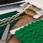 A collection of various rickrack trims in colors including white, gold, black, green, and grey are displayed on cardboard spools. A pair of scissors and some pins with yellow heads are also present on a wooden surface nearby.