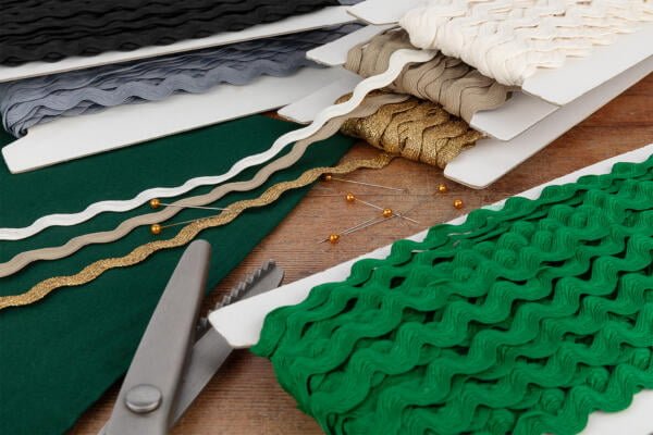 A collection of various rickrack trims in colors including white, gold, black, green, and grey are displayed on cardboard spools. A pair of scissors and some pins with yellow heads are also present on a wooden surface nearby.