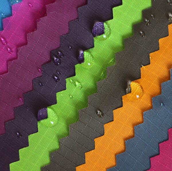 Close-up of colorful, zigzag-patterned fabric swatches with water droplets. The fabrics are arranged in diagonal stripes and feature bright colors, including pink, purple, green, black, orange, and blue. The droplets highlight the material's texture.