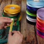 Two hands measure a green ribbon using a yellow measuring tape on a green cutting mat, next to a pair of scissors. Multiple colorful ribbon rolls, including shades of yellow, pink, red, blue, purple, black, and green, are stacked nearby on a wooden surface.