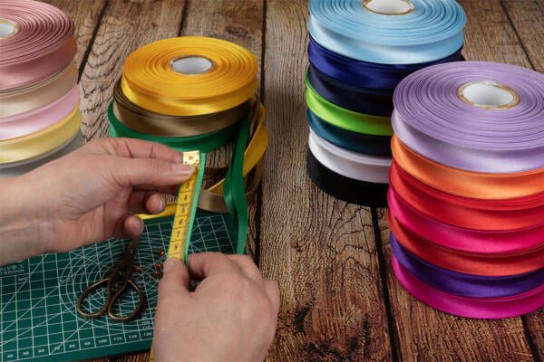 Two hands measure a green ribbon using a yellow measuring tape on a green cutting mat, next to a pair of scissors. Multiple colorful ribbon rolls, including shades of yellow, pink, red, blue, purple, black, and green, are stacked nearby on a wooden surface.