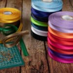 A wooden table with colorful spools of ribbon stacked on the right side. A green cutting mat with orange pins and loose green and brown ribbon rests on the left side. The ribbons are in various colors including blue, yellow, green, and red.