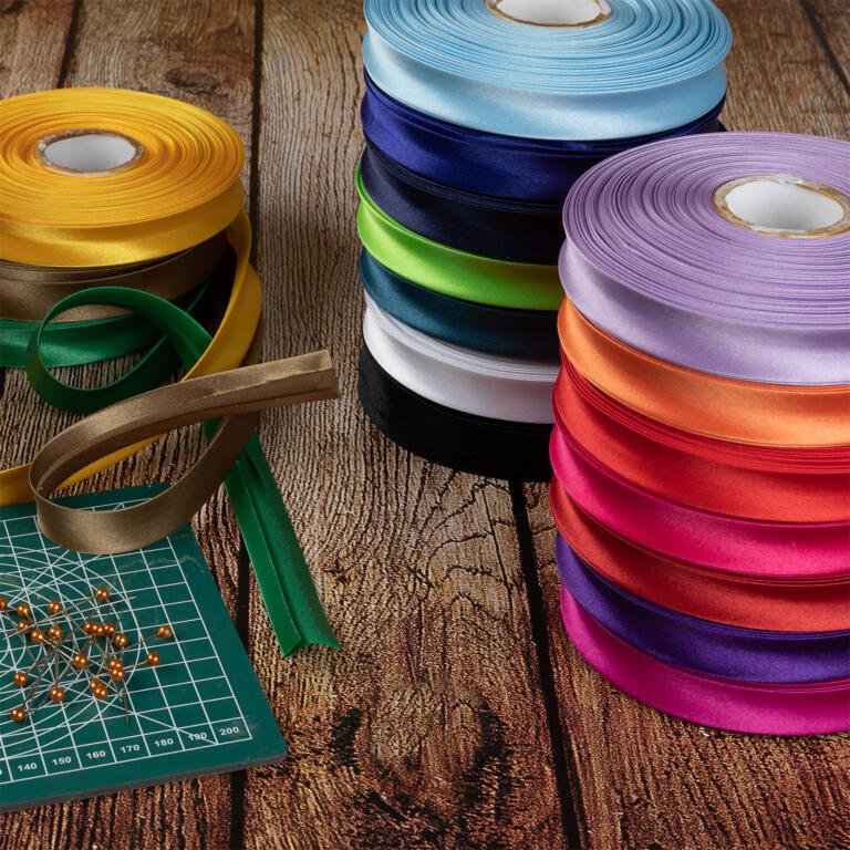 A wooden table with colorful spools of ribbon stacked on the right side. A green cutting mat with orange pins and loose green and brown ribbon rests on the left side. The ribbons are in various colors including blue, yellow, green, and red.