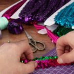 Hands arranging shiny sequins in pink, green, and purple on a crafting mat. Scissors, spools of thread, and colorful pins are also visible on the table, suggesting a crafting project in progress.