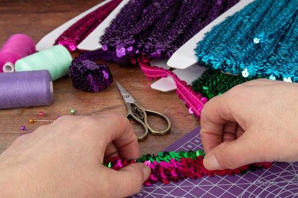 Hands arranging shiny sequins in pink, green, and purple on a crafting mat. Scissors, spools of thread, and colorful pins are also visible on the table, suggesting a crafting project in progress.