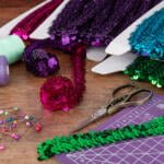 A wooden table with colorful sequined trims, spools of thread, sewing pins, and a pair of scissors. There is also a cutting mat with grid lines and a craft knife. The sequined trims are in shades of purple, green, and pink.