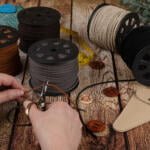 A person cutting brown braided leather cord with scissors. Spools of braided leather in various colors, a measuring tape, a ruler, buttons, a small leather pouch, and green plant leaves are arranged on a wooden surface.